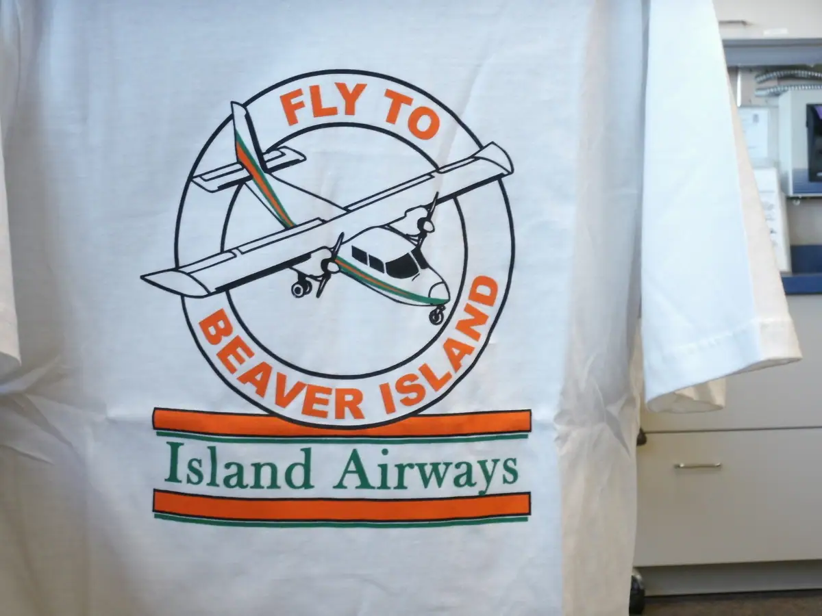 Photograph of a white T-shirt displaying an illustrated airplane logo and orange-and-green lettering.