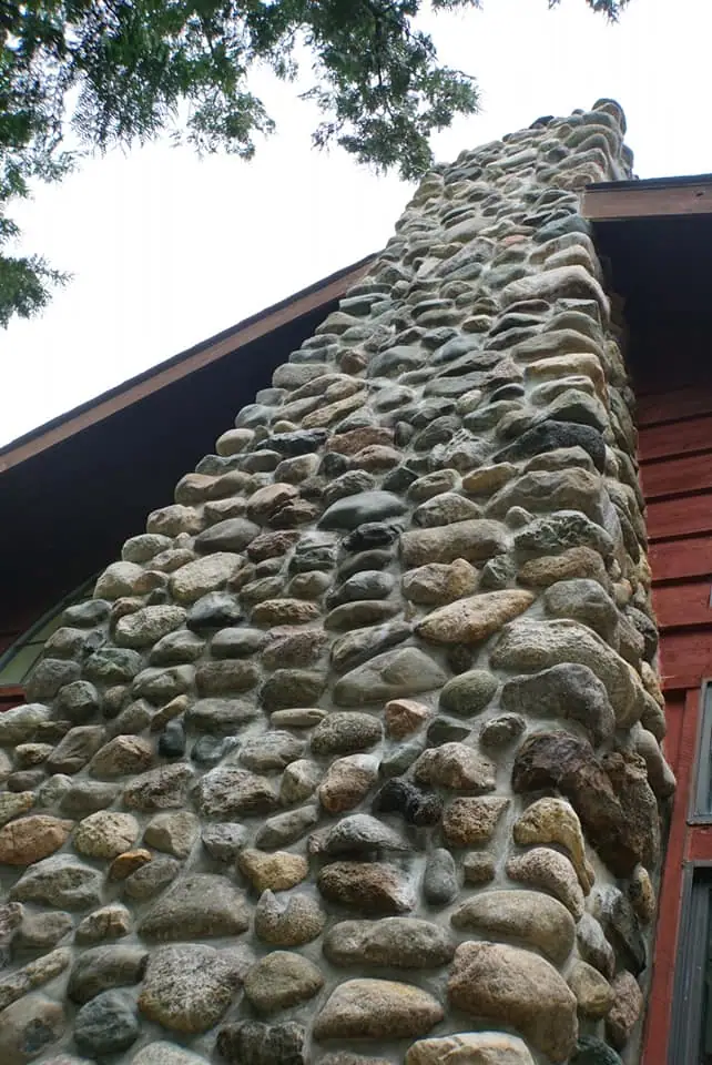Close-up view of a rounded-stone chimney rising beside the wooden exterior of a building.