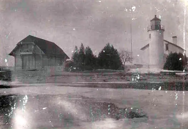 A cylindrical lighthouse and a large gabled building stand beside a tree line in a damaged monochrome photograph.