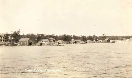 Sepia photograph of a small waterfront settlement with wooden buildings, trees, boats, and a water tower.