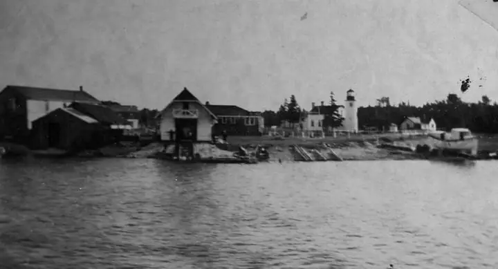 Black-and-white waterfront photograph showing a lighthouse, shoreline buildings, a pier, and rippling water.