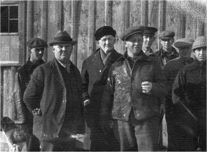Eight men pose outside a wooden building, with handwritten labels above several heads.