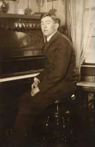 Sepia portrait of a seated man beside an ornate piano in a room with lace curtains.