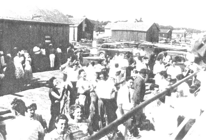 A dense crowd gathers on a pier beside wooden buildings and parked automobiles.