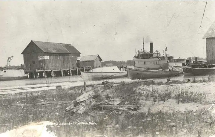 A weathered waterfront with wooden dock buildings, several boats, and a tugboat along the shore.