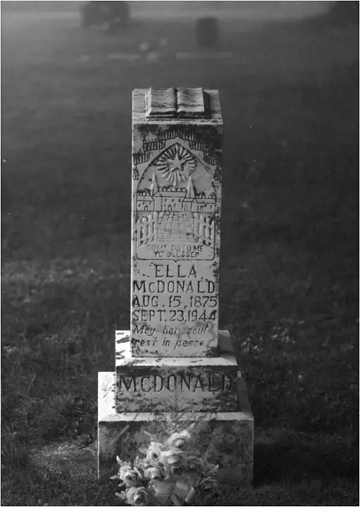 Black-and-white photograph of a decorated gravestone bearing a carved religious scene and floral arrangement.