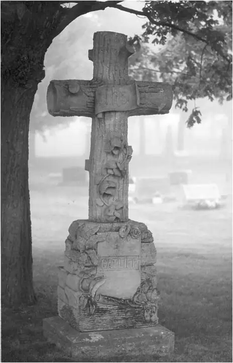A carved stone cross monument stands beneath a spreading tree, with blurred grave markers and vehicles behind it.