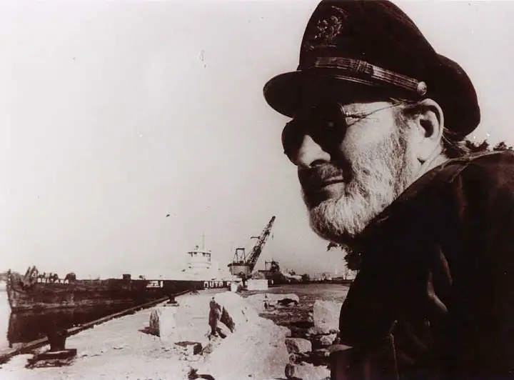 Bearded man in a peaked cap and sunglasses beside a docked freighter and crane.