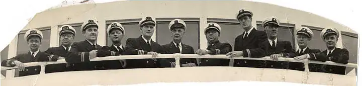Eleven uniformed men pose in a line behind a ship's railing.