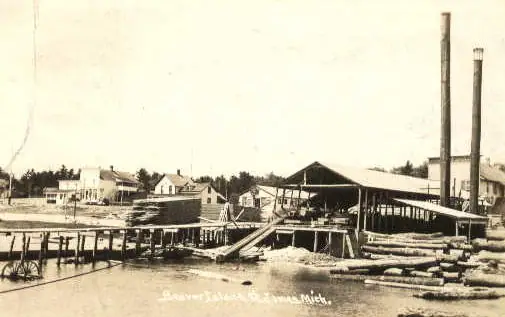 Sepia photograph of a waterfront lumber facility with docks, logs, buildings, and two tall smokestacks.