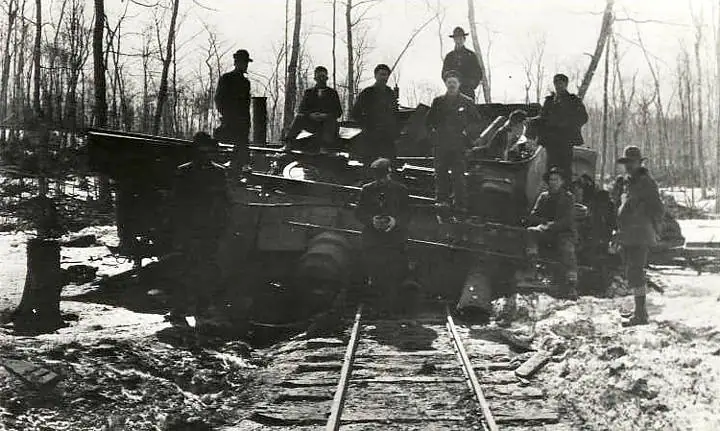 A group of people gathers around a rail-mounted machine on tracks in a snowy wooded setting.