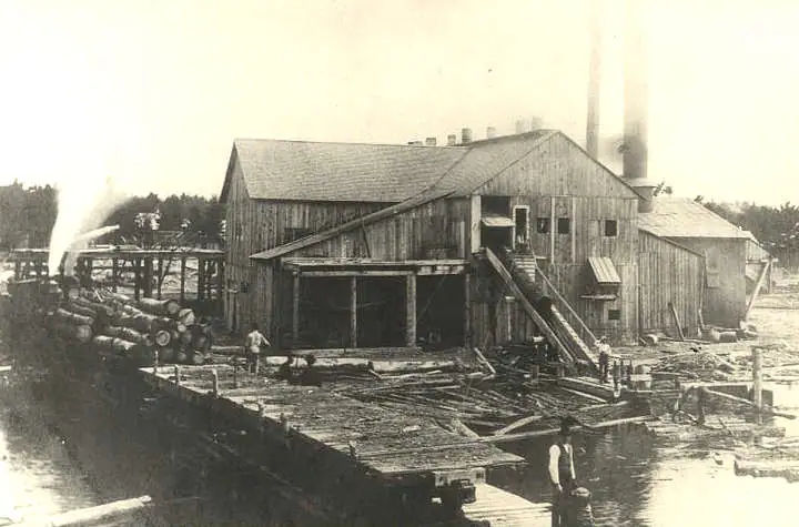 A weathered wooden lumber mill stands beside water, with logs, planks, workers, and smoke rising from its machinery.