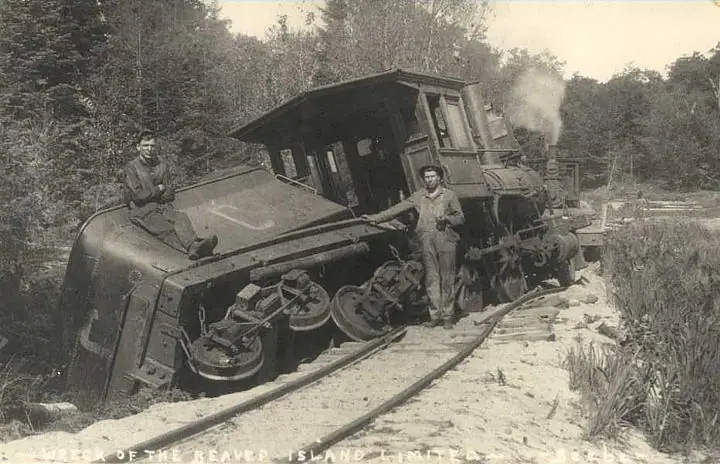 Derailed steam locomotive beside a wooded railway, with two men standing near the wreck.