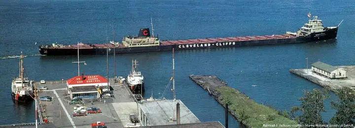 Aerial view of a bulk freighter passing a pier with smaller vessels, parked cars, and waterfront buildings.