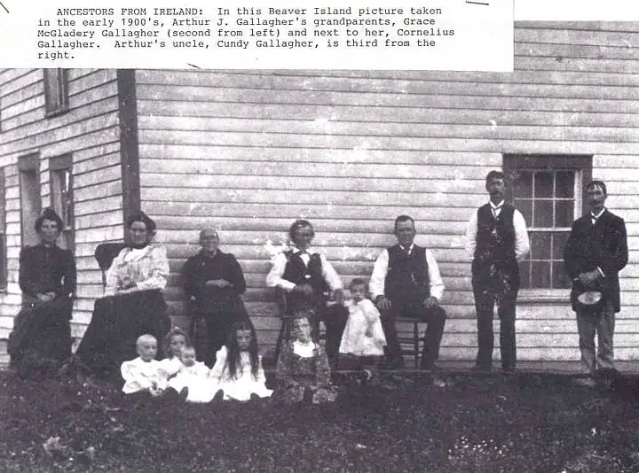 Black-and-white group portrait of adults and children posed outside a clapboard building.