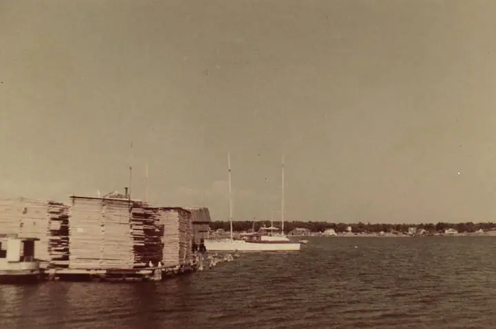 A lumber-loaded dock beside a white sailboat on calm water, with houses along the distant shoreline.