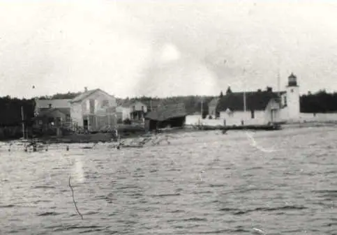 Black-and-white view across rippled water toward waterfront buildings and a lighthouse.