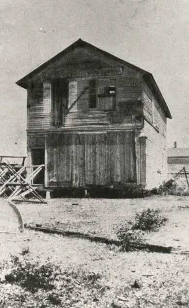 Black-and-white photograph of a weathered two-story wooden building with large plank doors and a gabled roof.