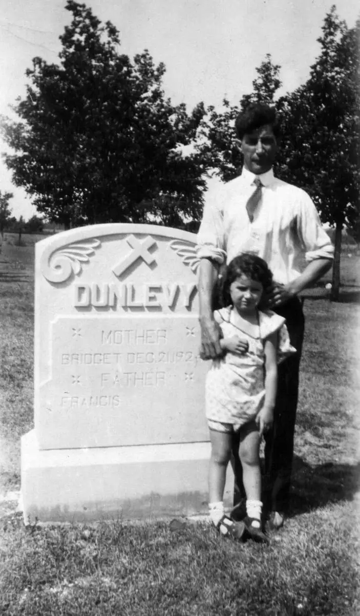 A man and young girl stand beside an ornate cemetery headstone bearing the name Dunlevy.