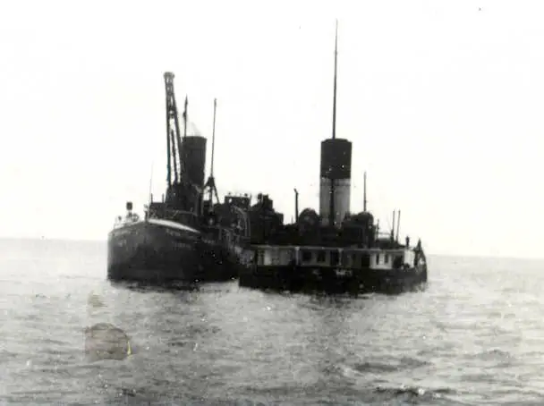 A large dark-hulled vessel with a tall smokestack and deck crane moves across rippled water.