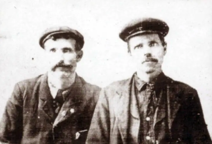 Black-and-white portrait of two mustached men in suits and caps standing side by side.