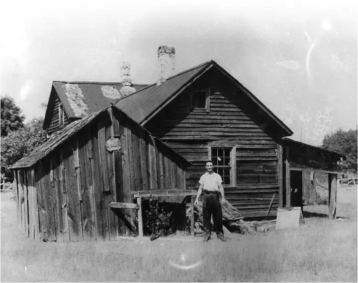 A man stands in front of weathered wooden buildings with gabled roofs and chimneys.
