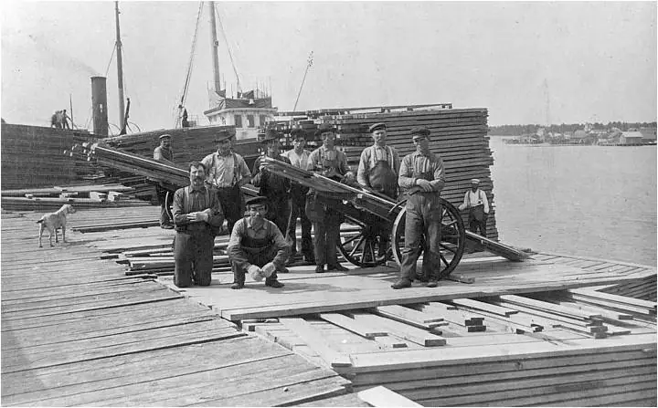 Men pose beside a lumber-loaded handcart on a wooden dock, with a ship, dog, and stacked planks behind them.