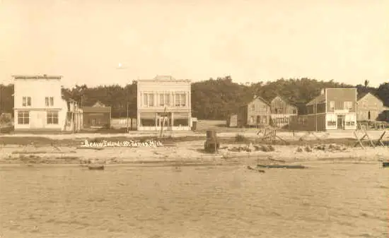 Sepia waterfront photograph showing a row of wooden buildings along a sandy shore.