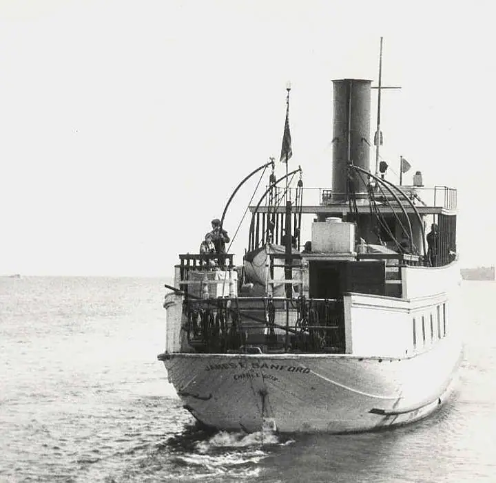 A black-and-white photograph of the steamer James E. Sanford viewed from astern on open water. Her transom reads JAMES E. SANFORD above CHARLEVOIX. Several figures stand at the after rail beneath a tall black funnel, with davits and a ship's boat on the deck above and an ensign flying.