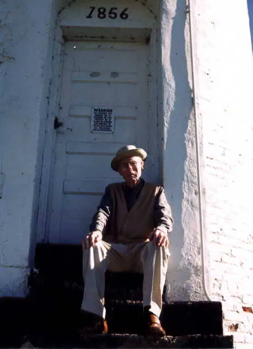 An older man sits on dark steps in front of a white-painted doorway marked 7856.
