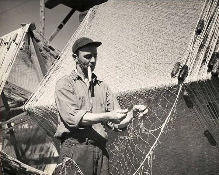 A man repairs a large fishing net while holding a long-stemmed pipe in his mouth.