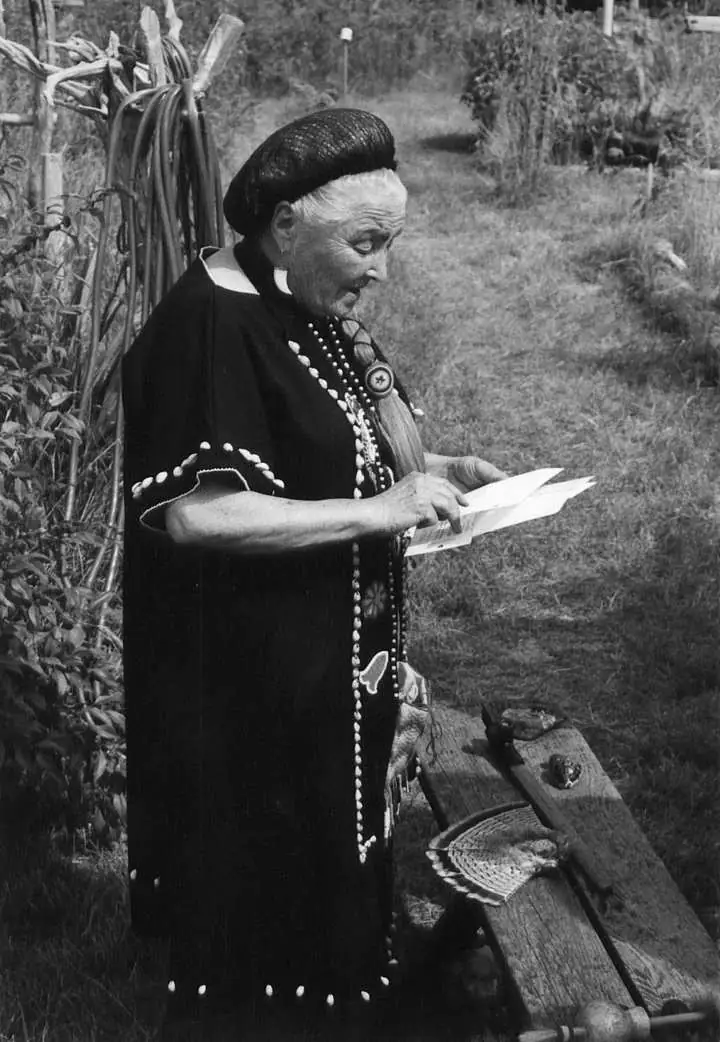 Black-and-white portrait of a woman in decorated clothing reading papers beside a wooden bench outdoors.