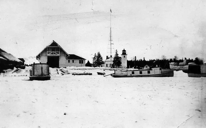 Black-and-white winter scene with a lighthouse, wooden buildings, and boats on a snow-covered shoreline.
