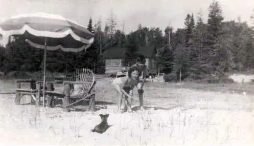 A woman crouches beside a dog on a sandy beach beneath a striped umbrella, with chairs and trees behind her.