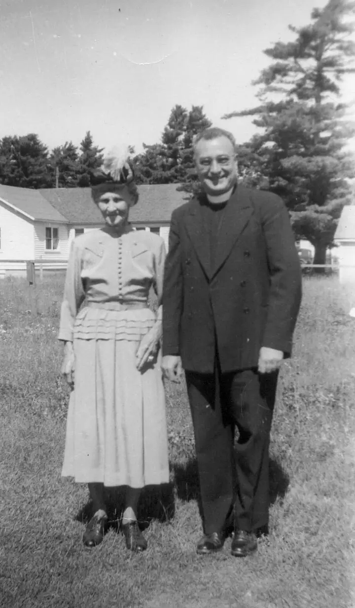 A woman in a feathered hat and a man in a dark suit pose in front of a house and trees.