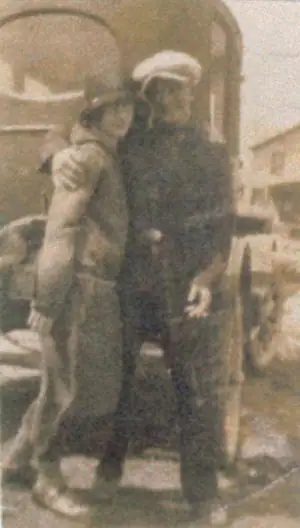 Two adults in hats stand together in front of a doorway in a sepia-toned photograph.