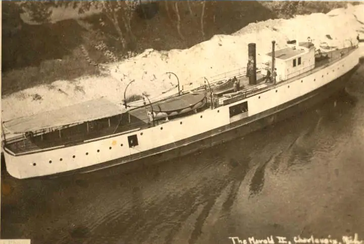 Sepia photograph of a long white ship with a smokestack moving past a wooded shoreline.