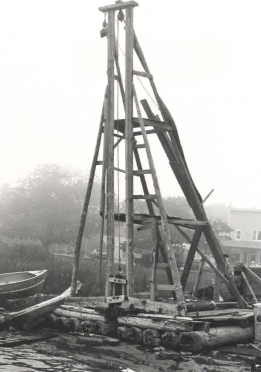 A tall wooden net-stake driver stands on a log raft beside small boats and a shoreline building.