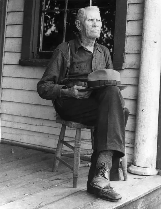 Black-and-white portrait of an older man seated on a porch, holding a broad-brimmed hat.
