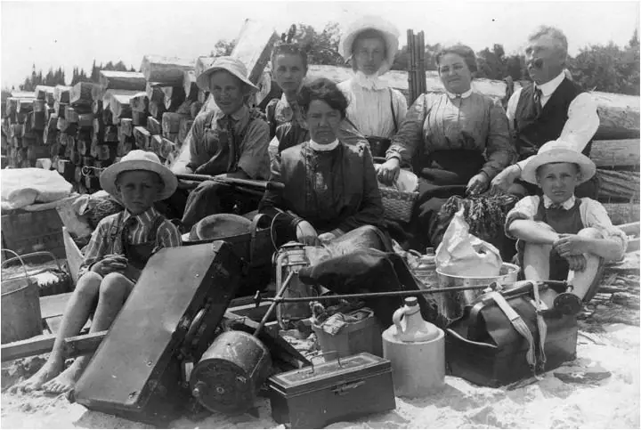 Seven people pose among equipment and belongings in front of a large stack of cut logs.