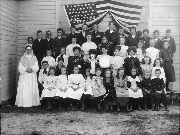 A large group of children and adults poses in front of a wooden building beneath a draped American flag.