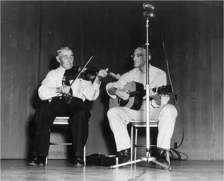Two seated musicians perform with a violin and acoustic guitar beside a microphone marked WUOM.
