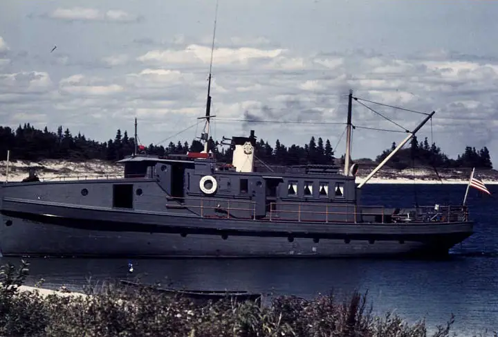 Dark patrol boat traveling on calm water beside a forested, sandy shoreline.
