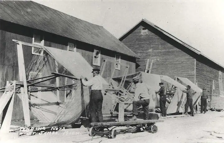 Several men work with large fishing nets on wooden frames outside two long wooden buildings.