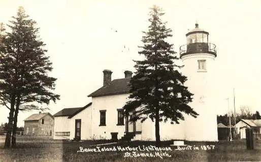 A white lighthouse and keeper’s dwelling stand among tall evergreen trees beside several small outbuildings.