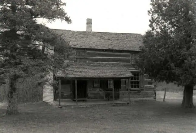 Black-and-white photograph of a log house with a covered porch, chimney, trees, and lawn.