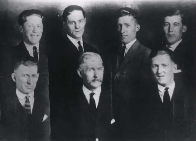 Black-and-white portrait of seven men in suits and ties arranged in two rows.