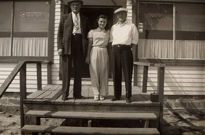 Three adults stand on a wooden porch in front of a building marked “COFFEE” and “BEER.”