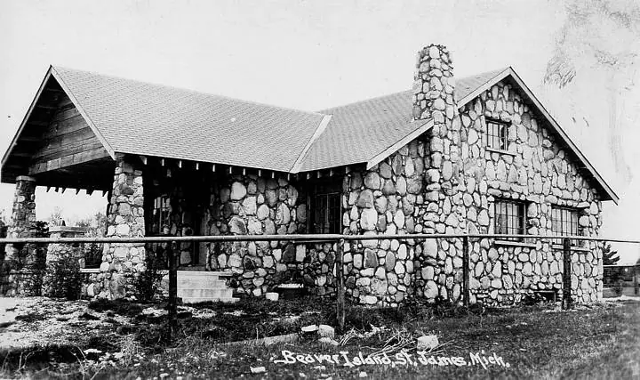 Black-and-white view of a large stone house with gabled roofs, a chimney, porch, and fence.