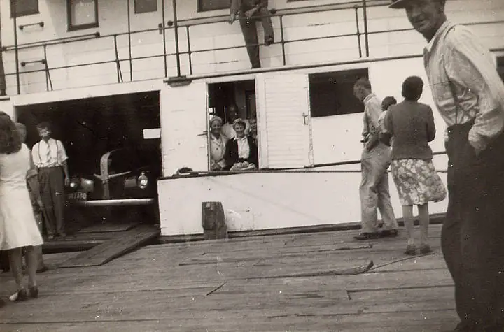People stand on the wooden deck of a ferry beside a vintage automobile.